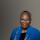 Senator Wanda Thomas Bernard, a black woman with short grey and black hair in her 60s wears grey blue glasses, a black shirt and a blue blazer with white flower pins on the left lapel. Around the blazer is a zebra-printed scarf with black and blue stripes. She is smiling.